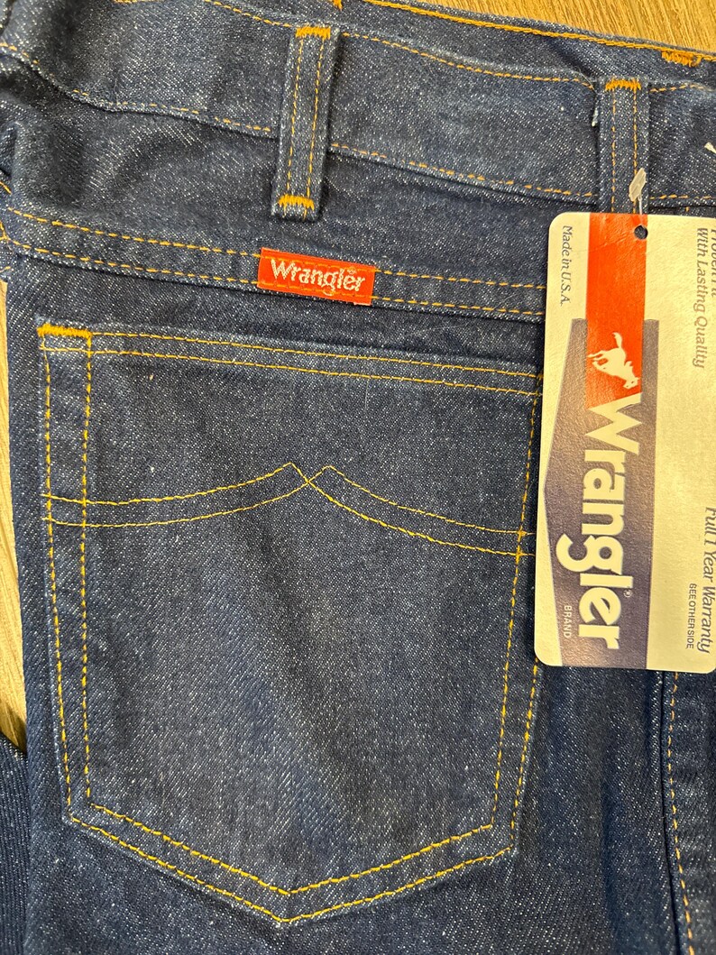 May include: Close-up of dark blue denim jeans with orange stitching. The back pocket features a classic design. A red Wrangler brand tag is visible. A paper tag with the Wrangler logo and text is attached.