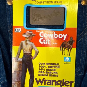 May include: A yellow and orange label with the text "The Original Cowboy Cut Jean" and a picture of a woman wearing blue jeans. The label also includes the text "ProRodeo Competition Jeans" and "Wrangler".