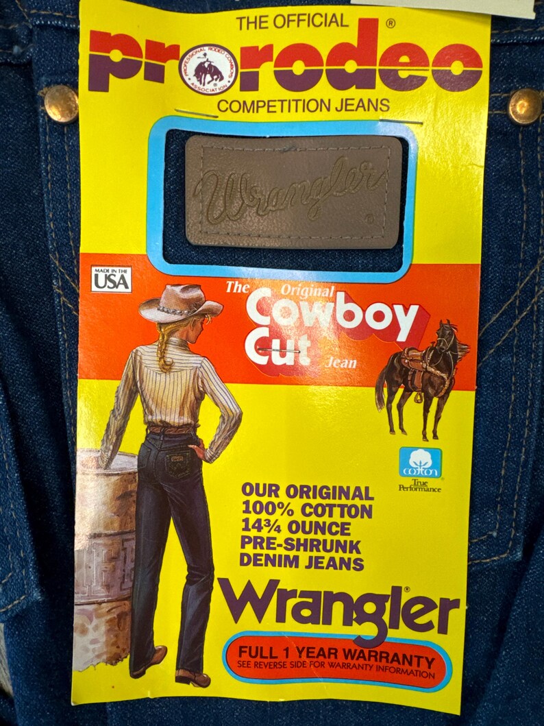 May include: A yellow and orange label with the text "The Original Cowboy Cut Jean" and a picture of a woman wearing blue jeans. The label also includes the text "ProRodeo Competition Jeans" and "Wrangler".