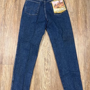 Puede incluir: Vaqueros Wrangler azul oscuro con un corte cl&aacute;sico, con un bolsillo trasero con un dise&ntilde;o de 'W' cosido. Los vaqueros tienen una pernera recta y una etiqueta con el logotipo de Wrangler. Los vaqueros se muestran sobre una superficie de madera.