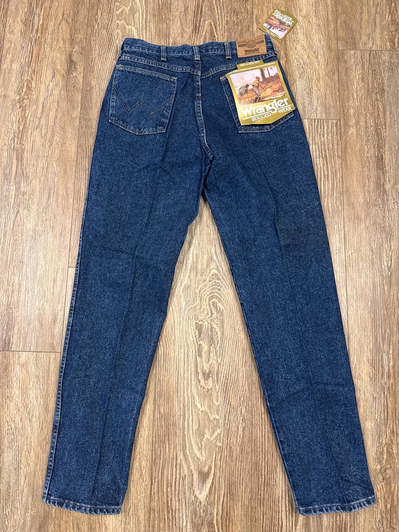Puede incluir: Vaqueros Wrangler azul oscuro con un corte cl&aacute;sico, con un bolsillo trasero con un dise&ntilde;o de 'W' cosido. Los vaqueros tienen una pernera recta y una etiqueta con el logotipo de Wrangler. Los vaqueros se muestran sobre una superficie de madera.
