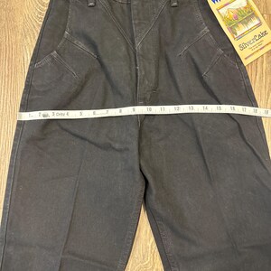 May include: Black Wrangler jeans with a high waist and decorative stitching. A measuring tape is laid across the front of the jeans. A tag that reads "Wrangler SilverLake" is attached to the jeans.