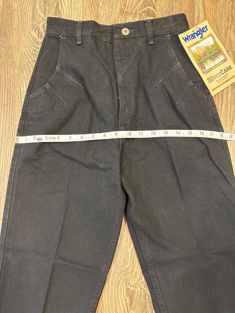 May include: Black Wrangler jeans with a high waist and decorative stitching. A measuring tape is laid across the front of the jeans. A tag that reads "Wrangler SilverLake" is attached to the jeans.
