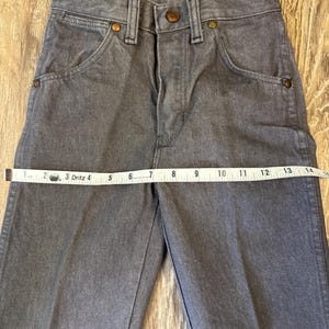 May include: Gray denim pants with a button and zipper closure, front pockets, and copper-colored rivets. A measuring tape shows the waist size. The pants are made of a durable denim material.