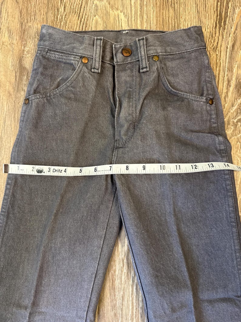 May include: Gray denim pants with a button and zipper closure, front pockets, and copper-colored rivets. A measuring tape shows the waist size. The pants are made of a durable denim material.