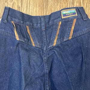 Puede incluir: Vaqueros de mezclilla azul oscuro con detalles de costura marr&oacute;n y una etiqueta Wrangler en la parte posterior de la cintura.