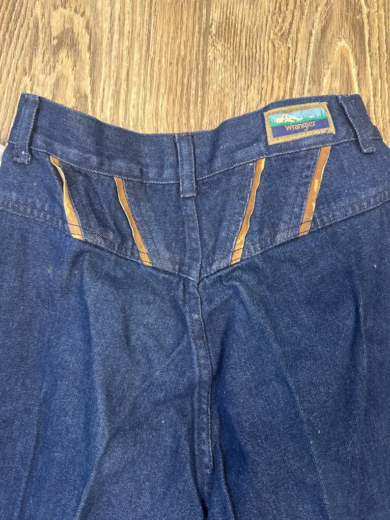 Puede incluir: Vaqueros de mezclilla azul oscuro con detalles de costura marr&oacute;n y una etiqueta Wrangler en la parte posterior de la cintura.