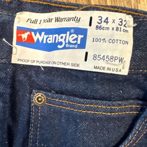 May include: Close-up of dark blue denim jeans with a Wrangler brand tag. The tag indicates a 34 x 32 size, equivalent to 86cm x 81cm, and is made of 100% cotton. The jeans have orange stitching and a metal button.