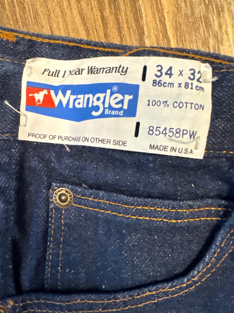 May include: Close-up of dark blue denim jeans with a Wrangler brand tag. The tag indicates a 34 x 32 size, equivalent to 86cm x 81cm, and is made of 100% cotton. The jeans have orange stitching and a metal button.