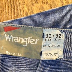May include: Close-up of a blue denim fabric with a Wrangler tag. The tag displays the brand name, size 32x32 (81cm x 81cm), and the text "100% COTTON". The tag also reads "Made in U.S.A."