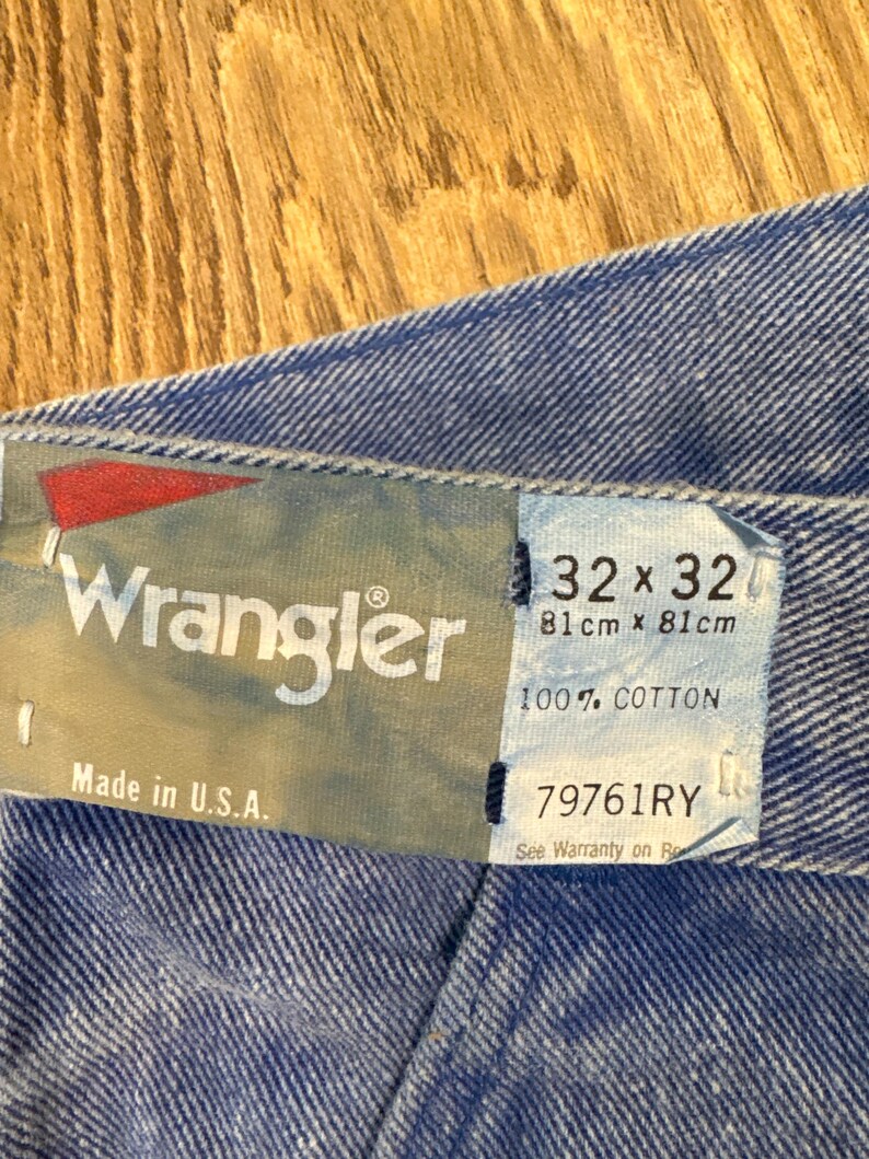 May include: Close-up of a blue denim fabric with a Wrangler tag. The tag displays the brand name, size 32x32 (81cm x 81cm), and the text "100% COTTON". The tag also reads "Made in U.S.A."