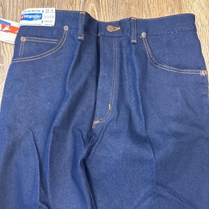 May include: Close-up of new, dark blue Wrangler jeans. The jeans have a button closure, front and back pockets, and are stitched with gold thread. A tag is attached to the jeans with the Wrangler logo and size information.