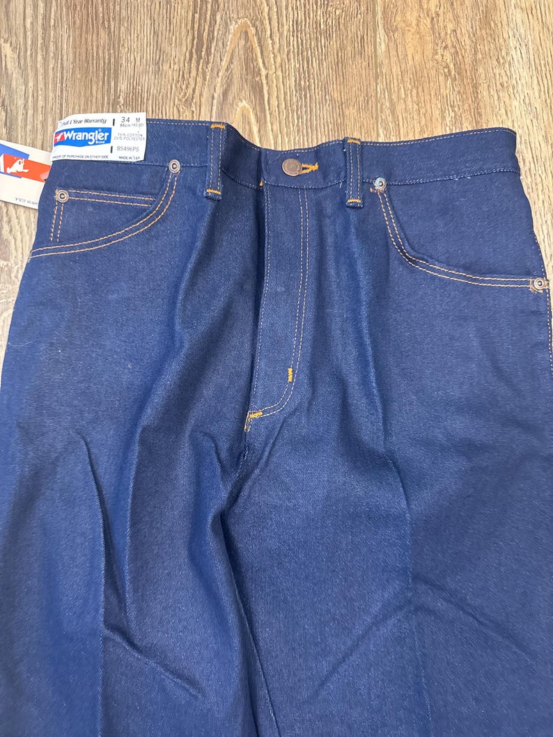 May include: Close-up of new, dark blue Wrangler jeans. The jeans have a button closure, front and back pockets, and are stitched with gold thread. A tag is attached to the jeans with the Wrangler logo and size information.