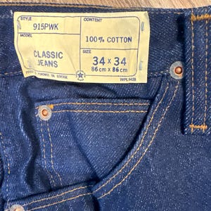May include: Close-up of dark blue Classic Jeans. The jeans have a label that reads "34 x 34" and "86 cm x 86 cm". The jeans are made of 100% cotton and have orange stitching.