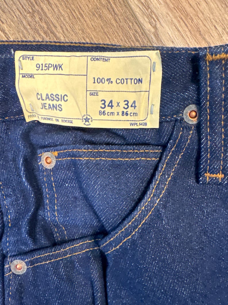 May include: Close-up of dark blue Classic Jeans. The jeans have a label that reads "34 x 34" and "86 cm x 86 cm". The jeans are made of 100% cotton and have orange stitching.