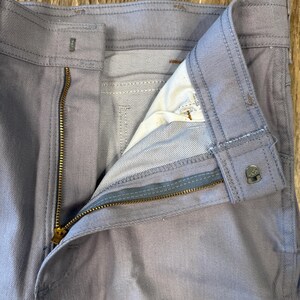 May include: Close-up of light blue denim pants with a partially unzipped brass zipper. The pants have a button closure and visible stitching details. The interior lining is visible, showing a lighter shade of blue.