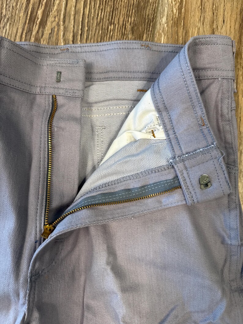 May include: Close-up of light blue denim pants with a partially unzipped brass zipper. The pants have a button closure and visible stitching details. The interior lining is visible, showing a lighter shade of blue.