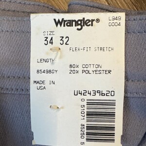 May include: Close-up of gray Wrangler Flex-Fit Stretch pants with a size 34/32 label. The tag indicates 80% cotton, 20% polyester, and "Made in USA". The label also displays a barcode and the Wrangler logo.