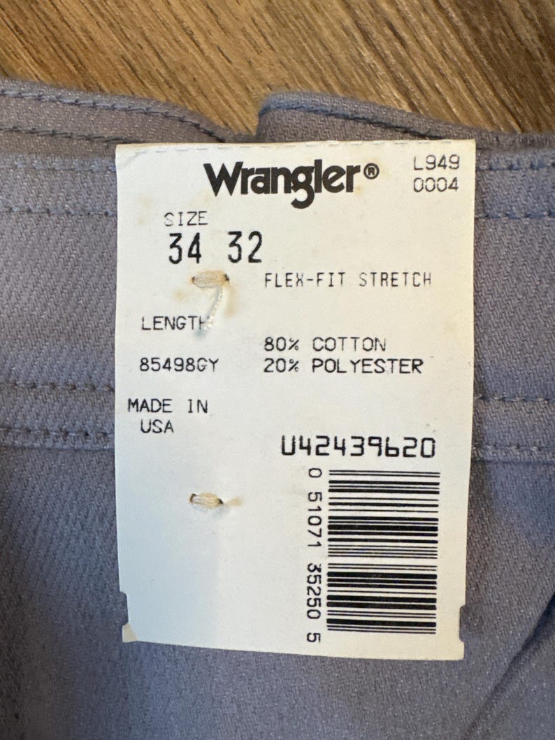 May include: Close-up of gray Wrangler Flex-Fit Stretch pants with a size 34/32 label. The tag indicates 80% cotton, 20% polyester, and "Made in USA". The label also displays a barcode and the Wrangler logo.