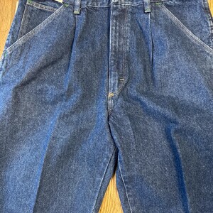 May include: Close-up of blue denim Wrangler jeans. The jeans have a classic design with a button closure, belt loops, and front pockets. The tag reads "Wrangler 34x32". The stitching is green.