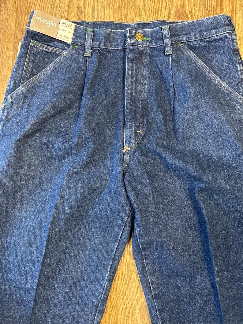 May include: Close-up of blue denim Wrangler jeans. The jeans have a classic design with a button closure, belt loops, and front pockets. The tag reads "Wrangler 34x32". The stitching is green.