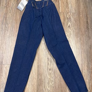 Puede incluir: Un par de pantalones vaqueros de mezclilla azul oscuro con cintura alta y pierna recta. Los vaqueros tienen un ribete de cuero marr&oacute;n en los bolsillos traseros.