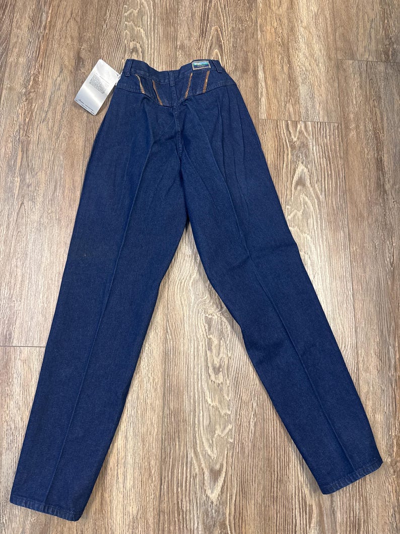 Puede incluir: Un par de pantalones vaqueros de mezclilla azul oscuro con cintura alta y pierna recta. Los vaqueros tienen un ribete de cuero marr&oacute;n en los bolsillos traseros.