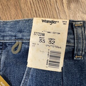 May include: Close-up of blue denim Wrangler jeans with a tag. The tag reads "Wrangler" and includes the waist size 84 cm and length 81 cm. The jeans are made of 100% cotton and have a "Antique Wash" label.