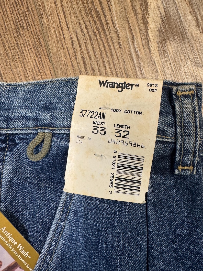 May include: Close-up of blue denim Wrangler jeans with a tag. The tag reads "Wrangler" and includes the waist size 84 cm and length 81 cm. The jeans are made of 100% cotton and have a "Antique Wash" label.