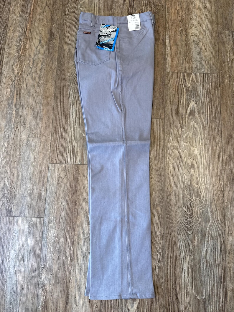 May include: Light blue denim pants with a straight leg. The pants have a classic five-pocket design and a Wrangler tag attached. The pants are laid flat on a wooden surface.