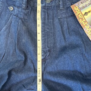 Puede incluir: Primer plano de pantalones vaqueros azul oscuro con cierre de bot&oacute;n y pliegues. Una cinta m&eacute;trica muestra una longitud de aproximadamente 38 cm. Los pantalones tienen una etiqueta Wrangler Riverlake.
