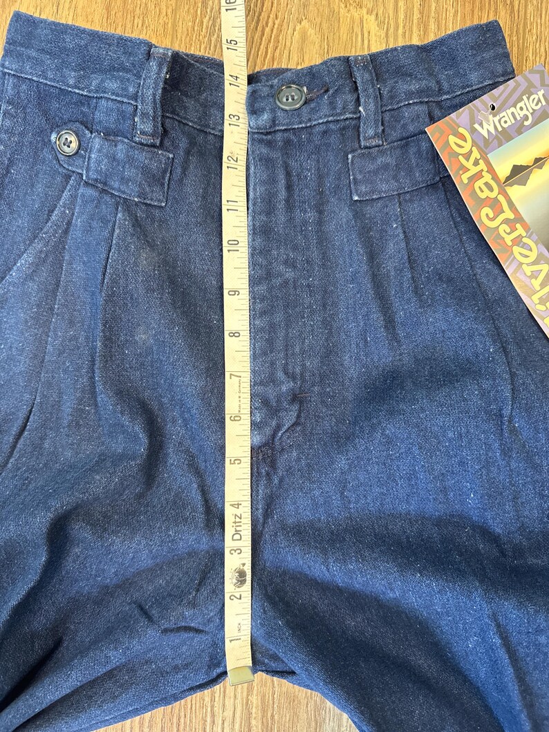Puede incluir: Primer plano de pantalones vaqueros azul oscuro con cierre de bot&oacute;n y pliegues. Una cinta m&eacute;trica muestra una longitud de aproximadamente 38 cm. Los pantalones tienen una etiqueta Wrangler Riverlake.