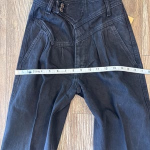 May include: Dark wash denim jeans with a high waist and pleated front. The jeans are measured with a tape measure, showing a waist measurement of approximately 40.6 cm across. The jeans have a button and zipper closure.