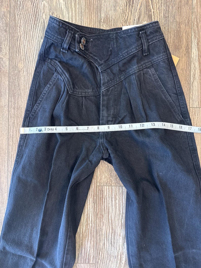 May include: Dark wash denim jeans with a high waist and pleated front. The jeans are measured with a tape measure, showing a waist measurement of approximately 40.6 cm across. The jeans have a button and zipper closure.