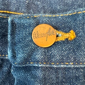 May include: A close-up of a copper-colored button with the word "Wrangler" embossed on it. The button is sewn onto a pair of blue denim jeans.