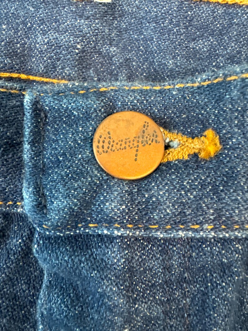 May include: A close-up of a copper-colored button with the word "Wrangler" embossed on it. The button is sewn onto a pair of blue denim jeans.