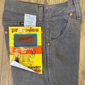 May include: Close-up of gray Wrangler Cowboy Cut jeans. The jeans have a classic five-pocket design with copper rivets and a brown leather patch. A yellow tag reads "pro rodeo" and "Cowboy Cut" with the Wrangler logo.