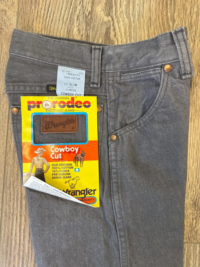 May include: Close-up of gray Wrangler Cowboy Cut jeans. The jeans have a classic five-pocket design with copper rivets and a brown leather patch. A yellow tag reads "pro rodeo" and "Cowboy Cut" with the Wrangler logo.