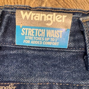 K&ouml;nnte beinhalten: Nahaufnahme einer blauen Jeans mit einem "Wrangler"-Etikett. Das Etikett lautet "STRETCH WAIST" und "STRETCHES UP TO 2.5 cm FOR ADDED COMFORT". Die Jeans hat einen Stretchbund f&uuml;r mehr Komfort.