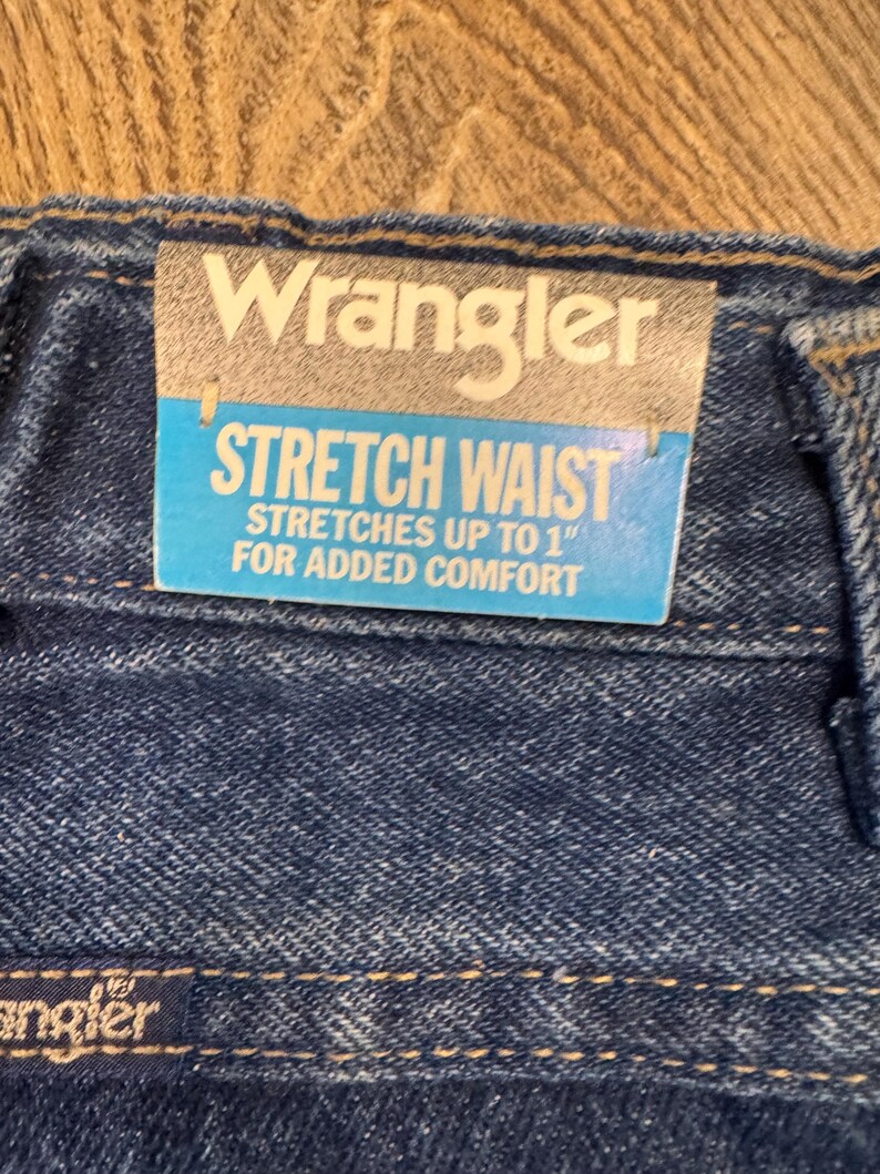 K&ouml;nnte beinhalten: Nahaufnahme einer blauen Jeans mit einem "Wrangler"-Etikett. Das Etikett lautet "STRETCH WAIST" und "STRETCHES UP TO 2.5 cm FOR ADDED COMFORT". Die Jeans hat einen Stretchbund f&uuml;r mehr Komfort.