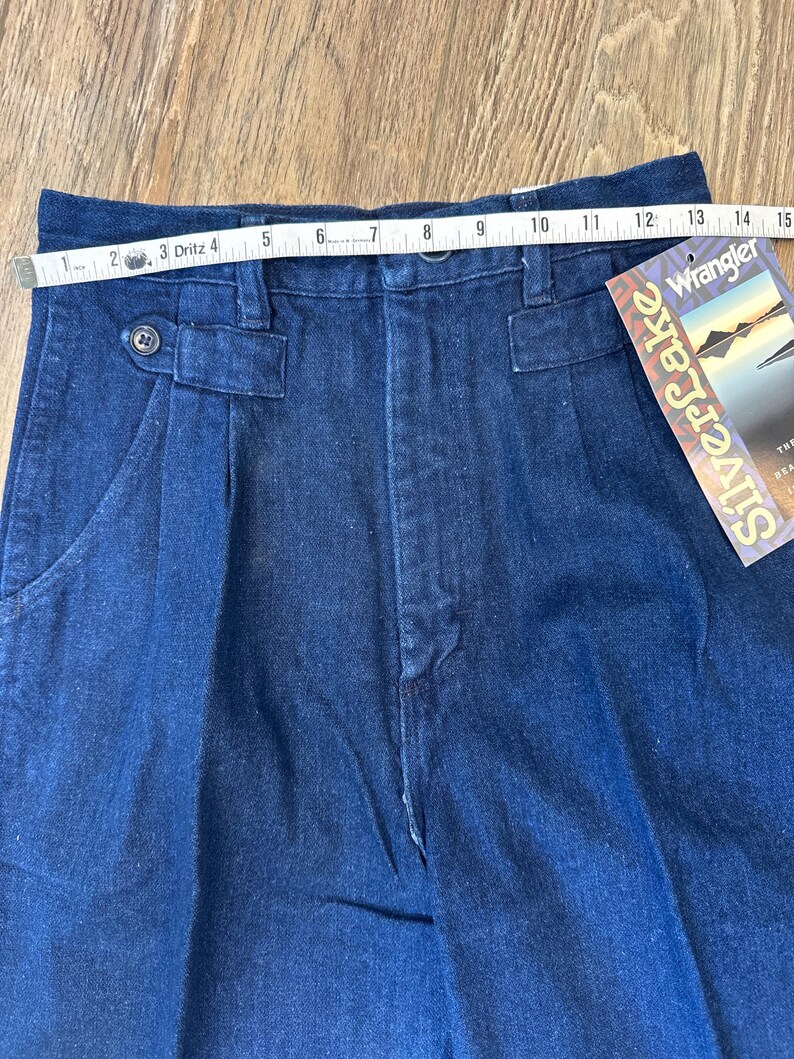 Puede incluir: Primer plano de pantalones vaqueros azul oscuro con cierre de bot&oacute;n y pliegues. Una cinta m&eacute;trica muestra una medida de cintura de aproximadamente 35.5 cm. Una etiqueta Wrangler Silverlake est&aacute; adjunta.