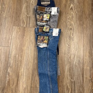 Puede incluir: Una pila de cuatro pares de pantalones vaqueros azules. El par superior es azul oscuro con el texto "American" y "Wrangler" en la etiqueta. El segundo par es azul claro con el texto "Wrangler" y "Rugged" en la etiqueta. El tercer par es de color gris claro con el texto "Wrangler" y "Rugged" en la etiqueta. El par inferior es de color azul medio con el texto "Wrangler" y "Classic" en la etiqueta.