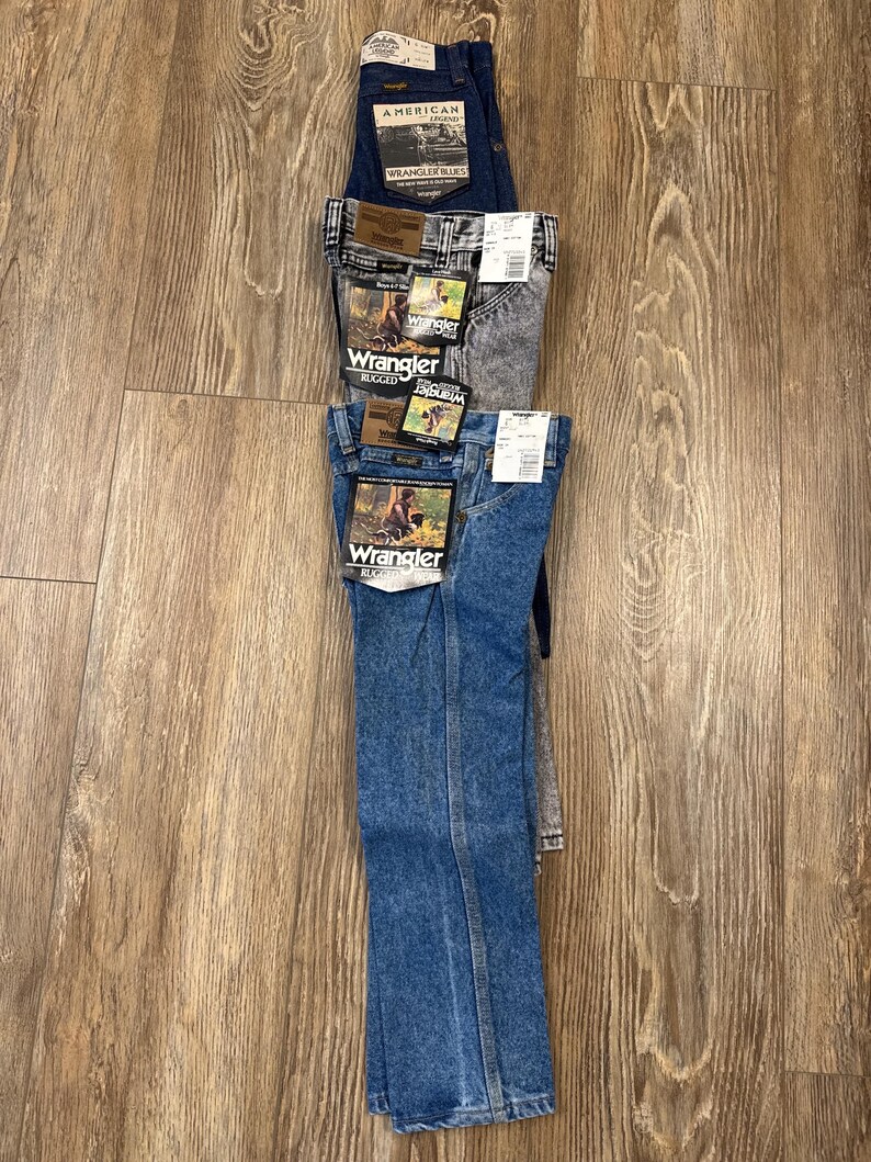 Puede incluir: Una pila de cuatro pares de pantalones vaqueros azules. El par superior es azul oscuro con el texto "American" y "Wrangler" en la etiqueta. El segundo par es azul claro con el texto "Wrangler" y "Rugged" en la etiqueta. El tercer par es de color gris claro con el texto "Wrangler" y "Rugged" en la etiqueta. El par inferior es de color azul medio con el texto "Wrangler" y "Classic" en la etiqueta.