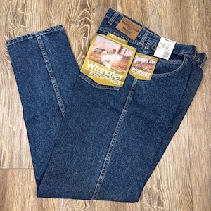 Puede incluir: Vaqueros Wrangler Rugged Wear de mezclilla azul con etiquetas. Los vaqueros est&aacute;n doblados y colocados sobre una superficie de madera. Las etiquetas presentan el logotipo de Wrangler y una imagen de un vaquero a caballo. Los vaqueros tienen un corte relajado.