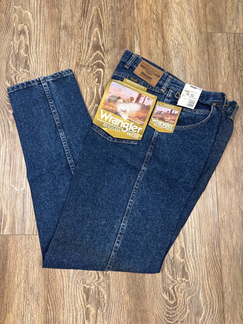 Puede incluir: Vaqueros Wrangler Rugged Wear de mezclilla azul con etiquetas. Los vaqueros est&aacute;n doblados y colocados sobre una superficie de madera. Las etiquetas presentan el logotipo de Wrangler y una imagen de un vaquero a caballo. Los vaqueros tienen un corte relajado.