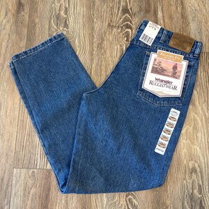 May include: A pair of blue denim jeans with a brown leather Wrangler Rugged Wear label. The label has the text "Relaxed Fit" and a picture of a cowboy riding a horse.