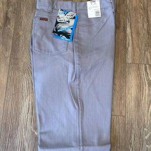 May include: Light gray Wrangler stretch jeans with a classic fit. The jeans have a straight leg, a button and zipper closure, and pockets. A Wrangler tag is attached to the back pocket. The jeans are displayed on a wooden surface.