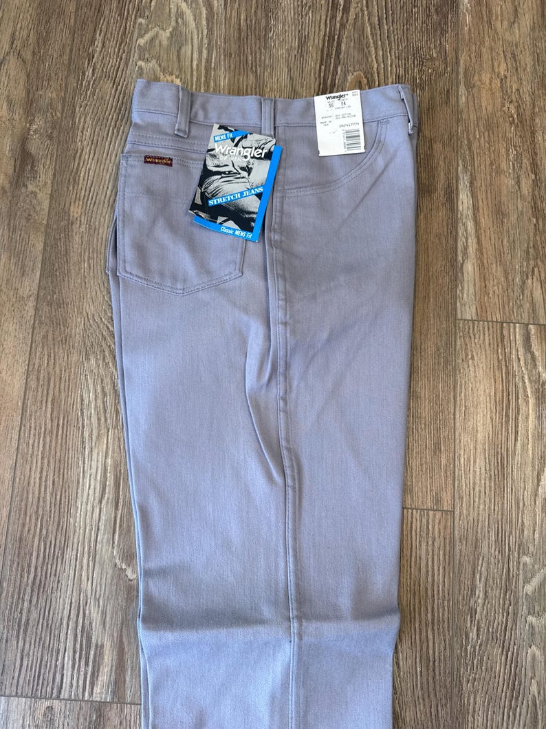May include: Light gray Wrangler stretch jeans with a classic fit. The jeans have a straight leg, a button and zipper closure, and pockets. A Wrangler tag is attached to the back pocket. The jeans are displayed on a wooden surface.
