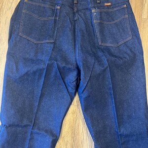 May include: Dark blue denim jeans with orange stitching and two back pockets. The jeans have a classic fit and a Wrangler tag attached. The jeans are folded and displayed on a wooden surface.