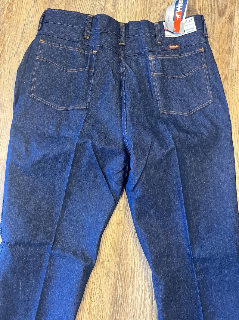 May include: Dark blue denim jeans with orange stitching and two back pockets. The jeans have a classic fit and a Wrangler tag attached. The jeans are folded and displayed on a wooden surface.